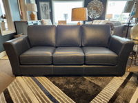 A Bayview Leather Sofa made by Leather Craft in contemporary black leather, featuring three cushions, is displayed in a living room setting, accented by lamps and decorative items in the background.