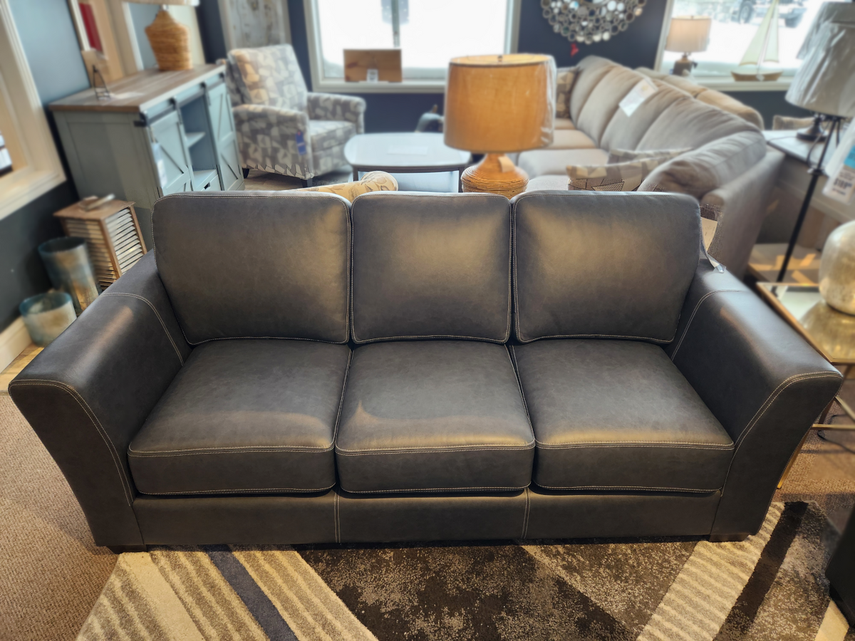 The Bayview Leather Sofa by Leather Craft, upholstered in dark blue leather and adorned with three cushions, is featured in a contemporary living room display alongside additional furniture and a stylish lamp.