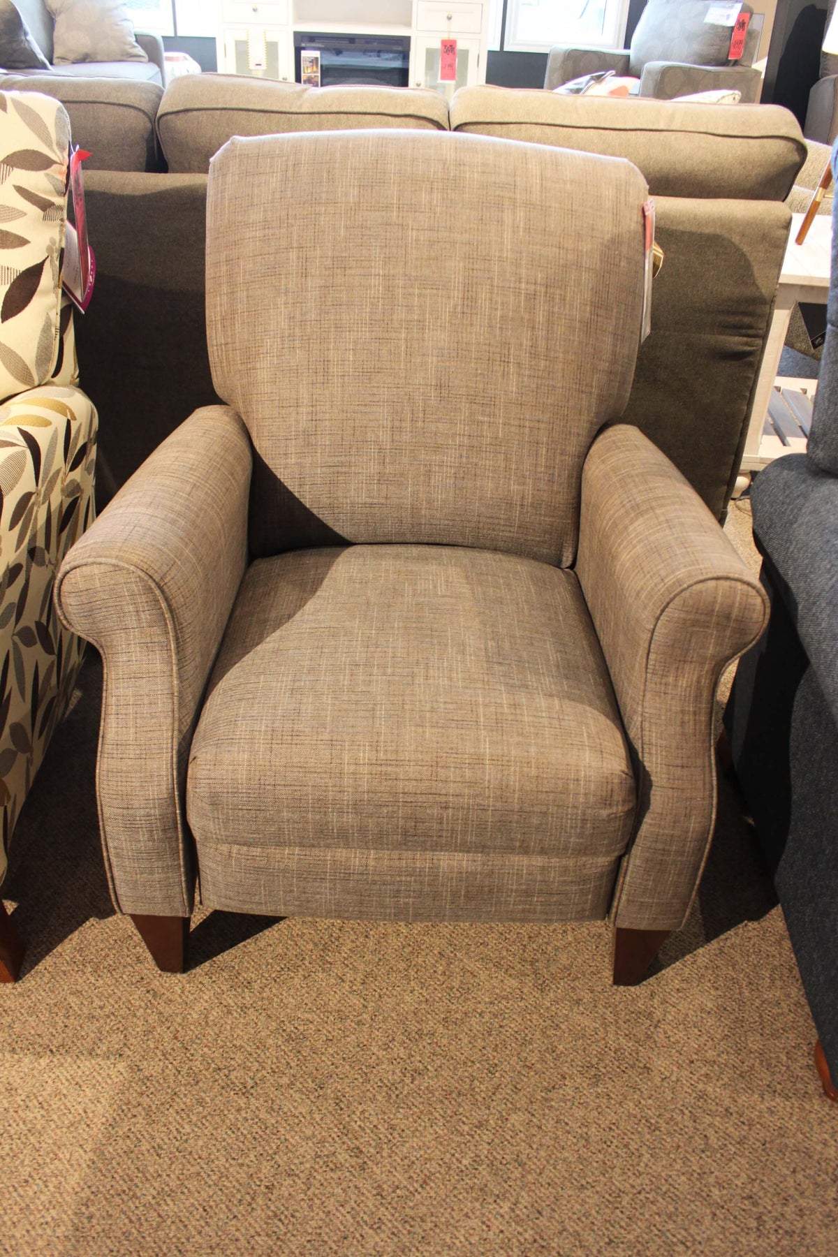 The La-z-boy 931 Charlotte High Leg Recliner, featuring a gray upholstered design with wooden legs, is showcased on a carpeted floor in a furniture store setting and offers customizable fabrics for the ideal personalized experience.