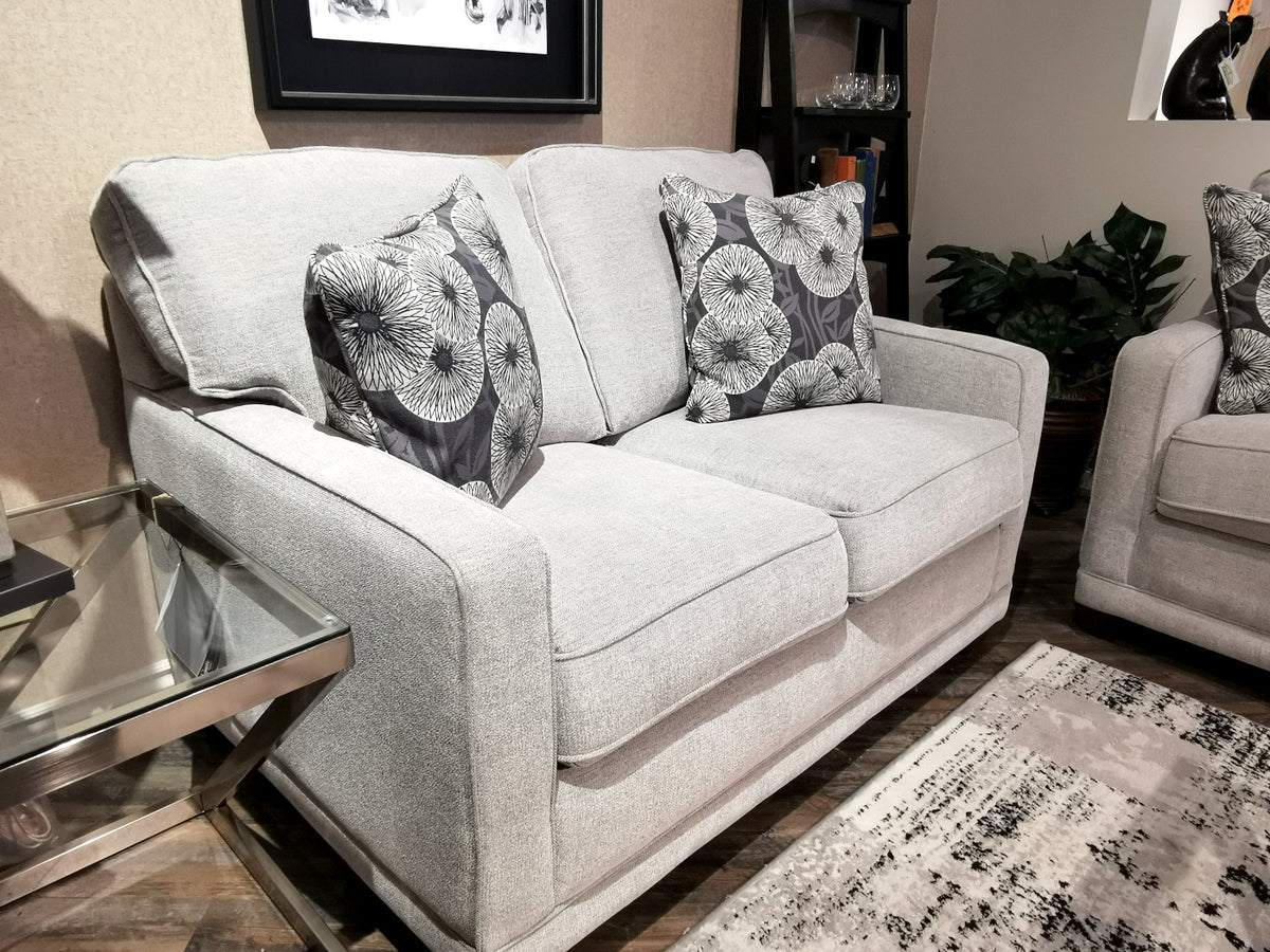 The 593 Kennedy Loveseat by La-Z-Boy, featuring gray upholstery with ComfortCore® cushions and patterned throw pillows, is positioned beside a glass side table in a cozy living room setting.