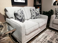 The 593 Kennedy Loveseat by La-Z-Boy, featuring gray upholstery with ComfortCore® cushions and patterned throw pillows, is positioned beside a glass side table in a cozy living room setting.