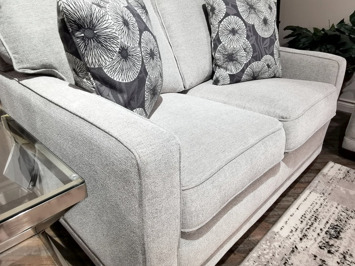 The 593 Kennedy Loveseat by La-Z-Boy in light gray showcases ComfortCore® cushions and includes two patterned pillows, positioned beside a glass side table. A plant is visible in the background, enhancing the cozy ambiance with your preferred fabric choice.