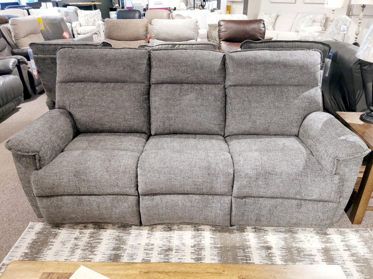 A gray 706 Jay Power with headrest three-seater sofa from La-Z-Boy exudes reclining comfort as it sits elegantly in a furniture store, poised on a patterned carpet with a wooden coffee table nearby.