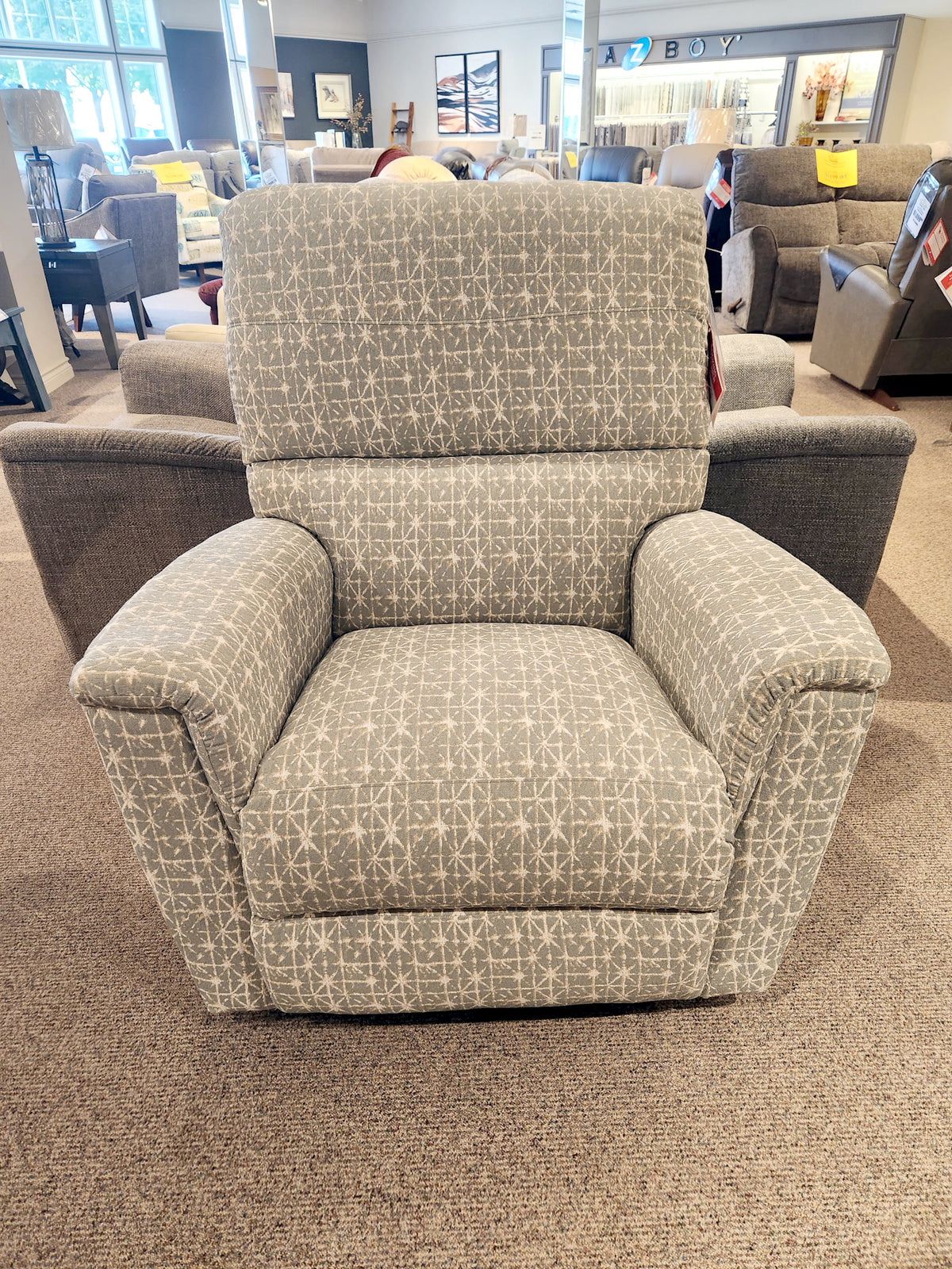 The 769 Ava Rocker Recliner by La-z-boy, featuring a gray patterned design, is showcased in a furniture store showroom, providing adjustable support and adding stylish elegance to any living space.