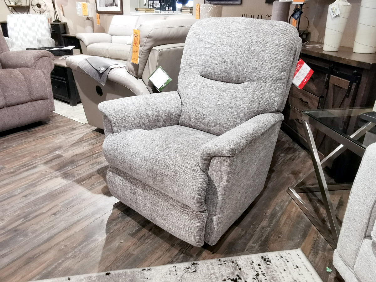 A 783 Aries Rocker Recliner from La-z-boy, upholstered in gray, takes center stage in a furniture store, surrounded by similar seating options. Discover this premium rocker recliner and explore its customizable fabric options for an unparalleled comfort experience.