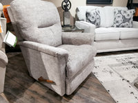 The La-z-boy 783 Aries Rocker Recliner, upholstered in gray and crafted from premium-grade fabrics, sits on a wooden floor next to a couch adorned with floral pillows and a patterned rug.