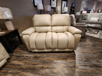 The La-Z-Boy 530 Greyson Reclining Loveseat, with its beige leather finish, is displayed in a showroom with wood flooring. It harmonizes with the surrounding furniture and provides a power recline option for maximum comfort.