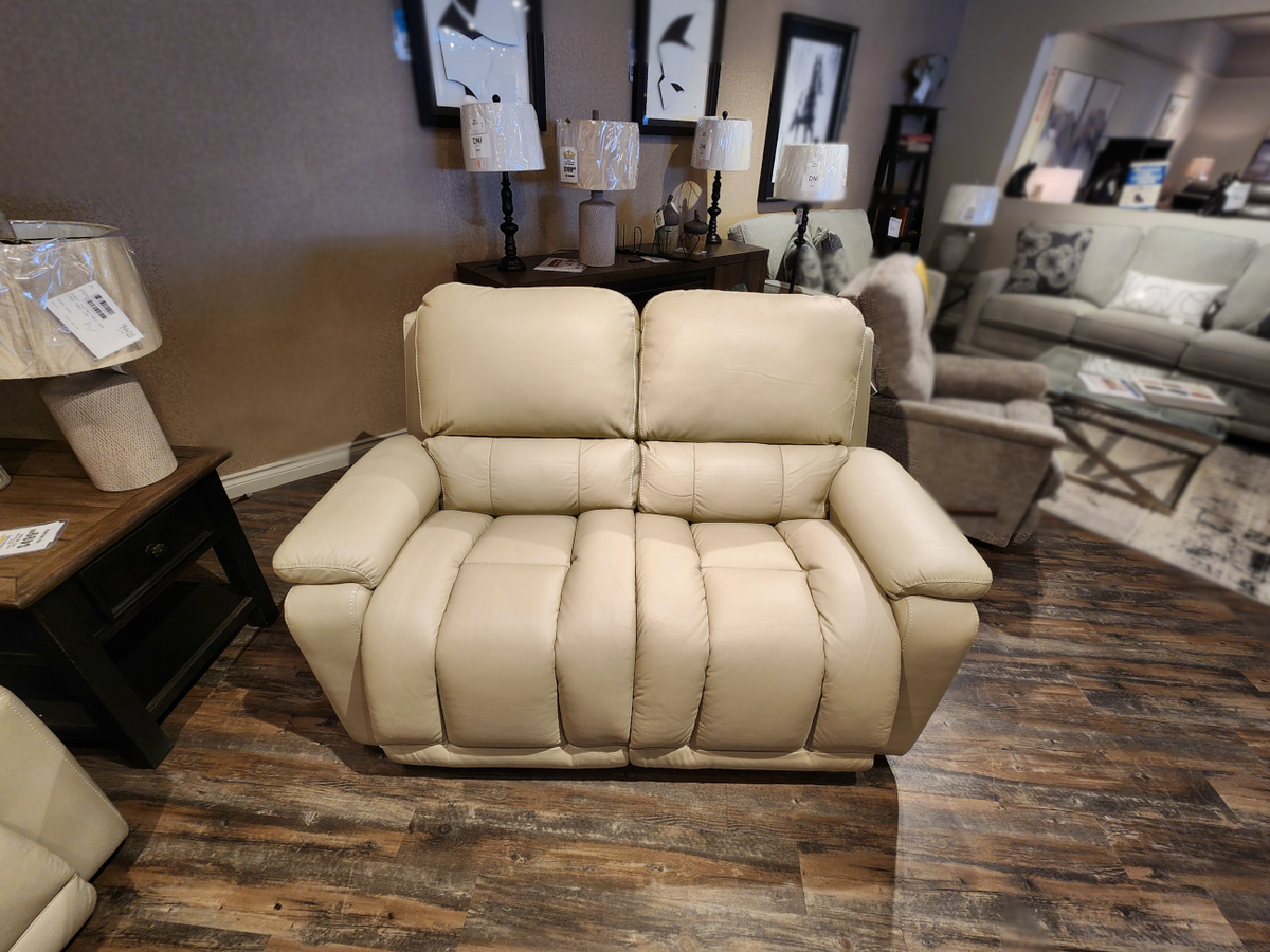 La-z-boy's 530 Greyson Reclining Loveseat, upholstered in beige leather, is displayed on a wooden floor amidst lamps and artwork in a furniture showroom, offering a power recline option for ultimate comfort.