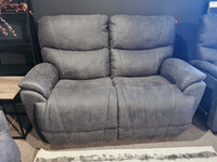 The 724 Trouper Loveseat by La-Z-Boy features a gray color with reclining bucket seats and stitching detail, positioned against a dark wall on a carpeted floor.