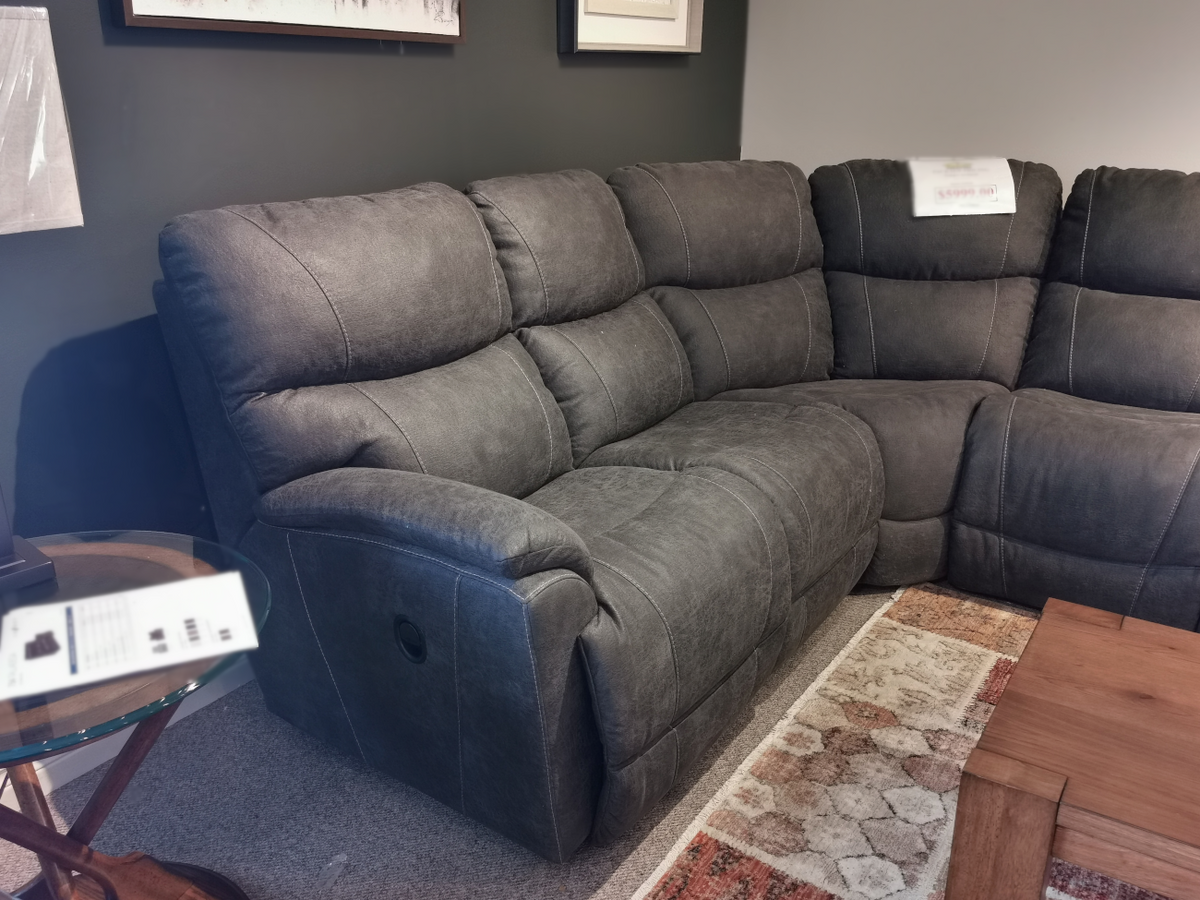 The La-z-boy 724 Trouper Reclining Corner Sectional in gray upholstery offers comfortable seating for three, situated on a carpeted floor beside a wooden side table and coffee table. Experience its comfort and elegance, all supported by our limited lifetime warranty for your peace of mind.