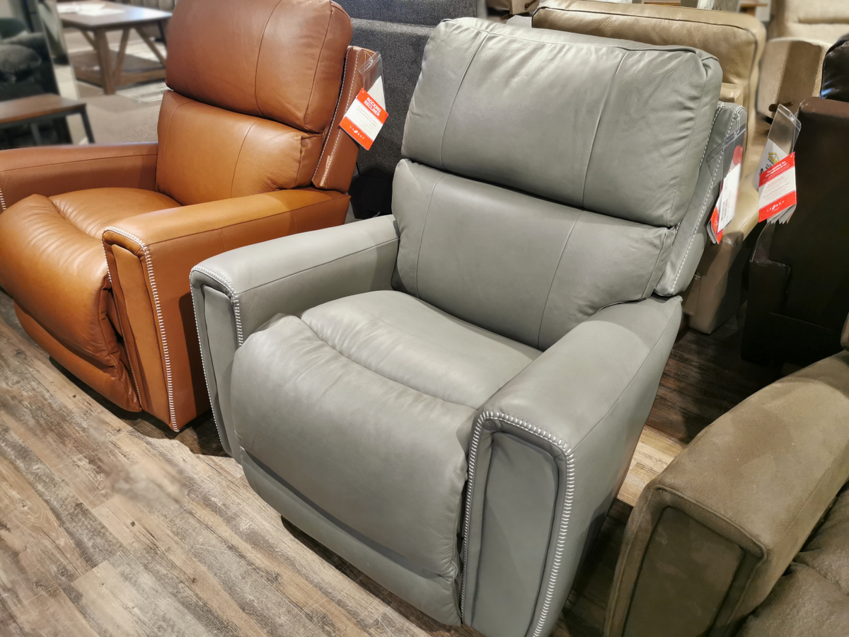 A gray recliner chair featuring modern comfort is showcased in a store, marked with its price. Next to it sits a brown leather La-Z-Boy 757 Apollo Rocker Recliner, both positioned on a wooden floor and inviting customers with their plush bucket seats.