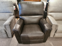 The La-z-boy 769 Ava Rocker Recliner, crafted in brown leather, is showcased between a gray and beige chair in a showroom setting, featuring adjustable support for enhanced comfort.