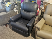 The 783 Aries Rocker Recliner by La-Z-Boy, crafted from luxurious black leather, is showcased in a furniture store. Among other chairs on a carpeted floor, it offers unmatched comfort and durability, making it the perfect choice for those looking for style and relaxation in a reclining rocker.