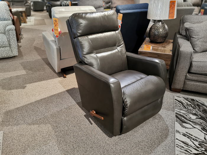 The 787 Lennon Leather Rocker Recliner by La-Z-Boy, featuring a dark leather finish and oversized comfort, takes center stage on the showroom floor. Encircled by other elegant furniture items, its tailored design is matched by a discreet price tag.