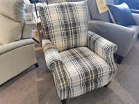 The 931 Charlotte High Leg Recliner by La-z-boy, featuring a plaid pattern in dark and light gray tones, is displayed in a furniture store. It is positioned between two other chairs to highlight its customizable fabric options for personalized style.