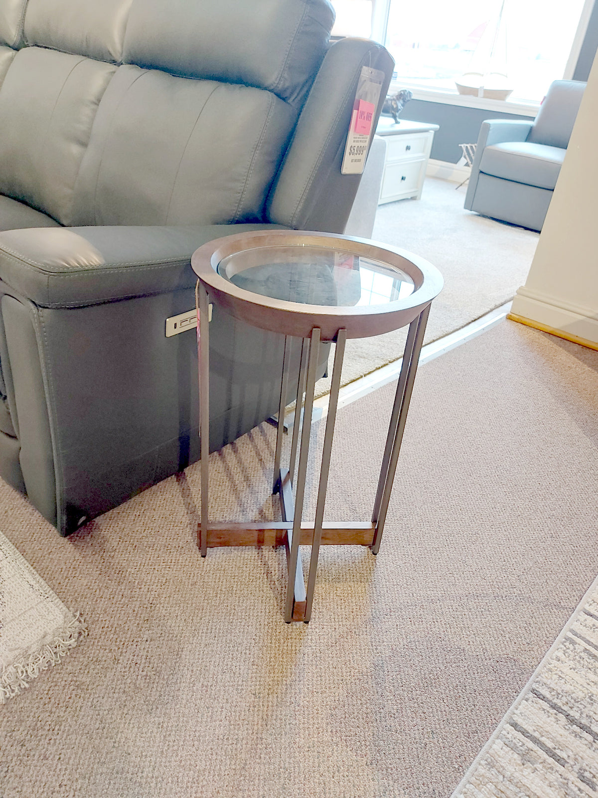 The Magnussen T5639 Elora Round Accent Table, showcasing a small round glass top and sleek art deco design, features a dark wooden frame with luxurious hickory veneers. It's ideally positioned beside a gray sofa on the carpeted floor in the living room.