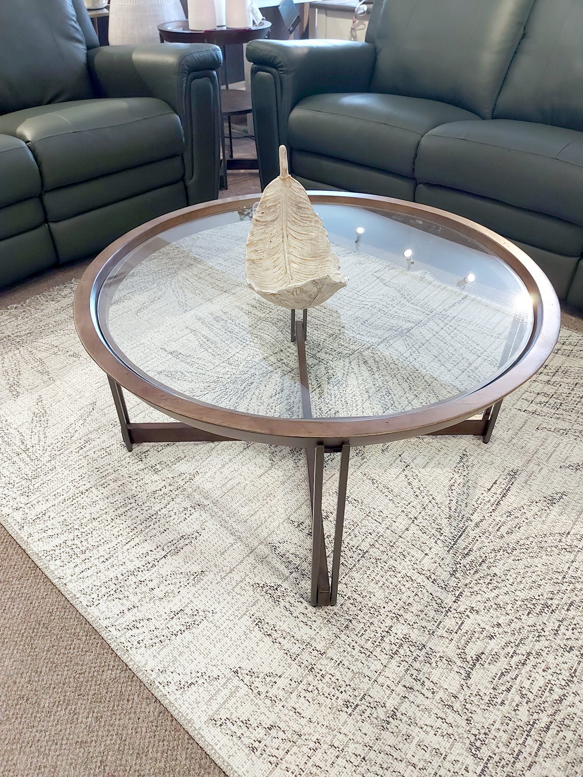 The T5639 Elora Round Cocktail Table by Magnussen, featuring a glass top and walnut-finished wooden frame, is placed on a patterned rug between two green sofas, with a leaf sculpture centerpiece that highlights a modern mix of materials.