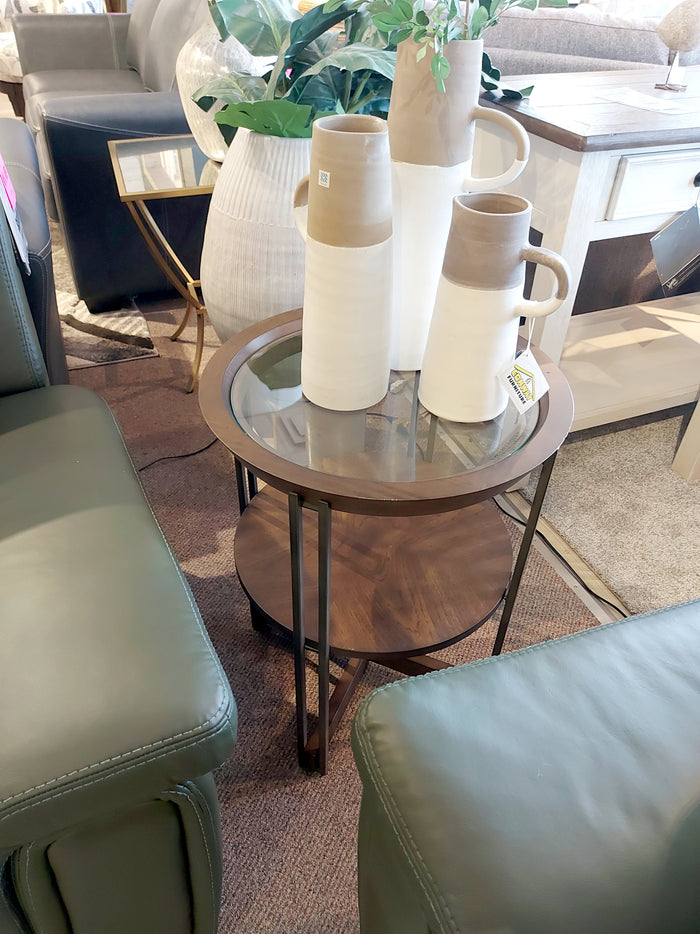 The T5639 Elora Round End Table by Magnussen is a round glass-top side table featuring dark wood and metal legs with hickory veneers. It includes three decorative vases and is complemented by green chairs, bringing an Art Deco flair to your space.