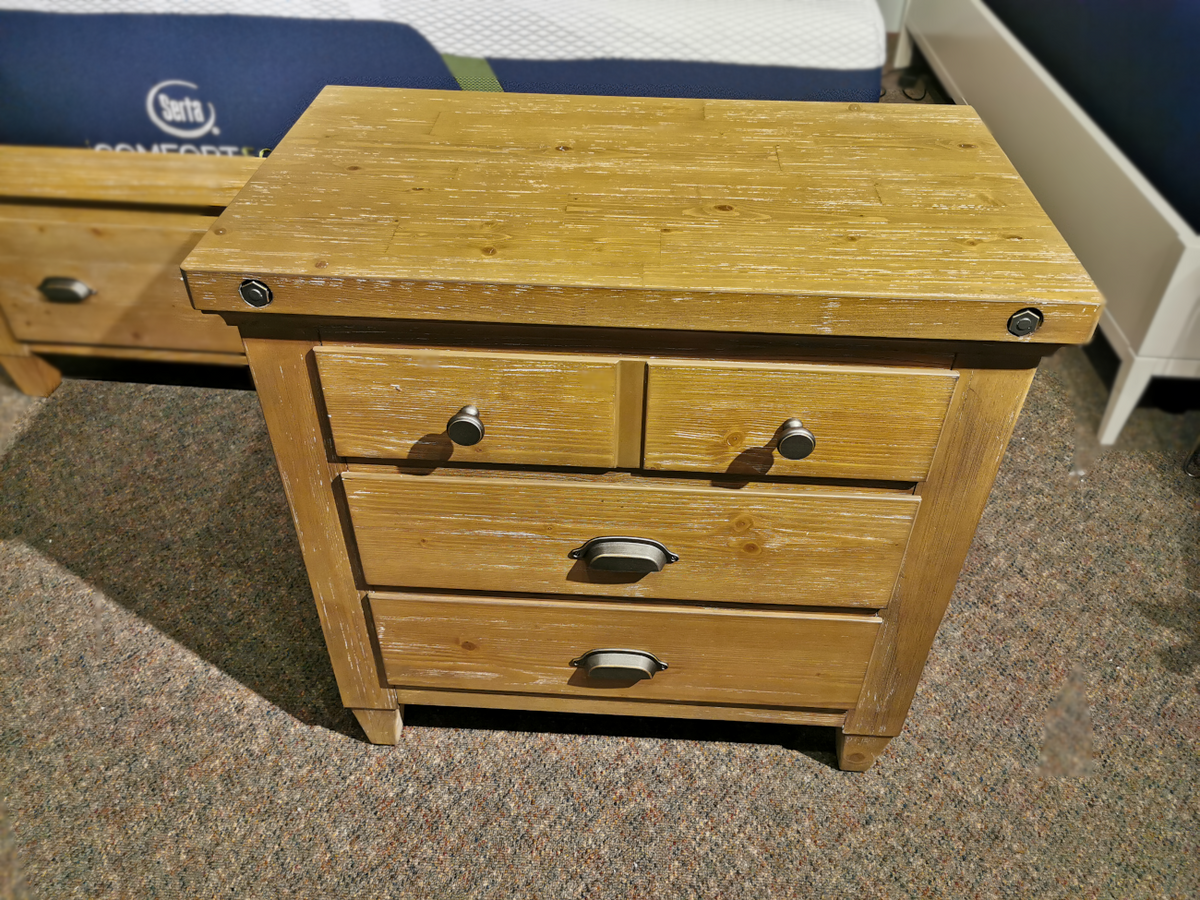 The Magnussen B5487 Lynnfield Nightstand, featuring a rustic design crafted from wirebrushed pine with a Weathered Fawn finish, boasts three drawers with metal handles and stands elegantly on a carpeted floor in a cozy room setting.