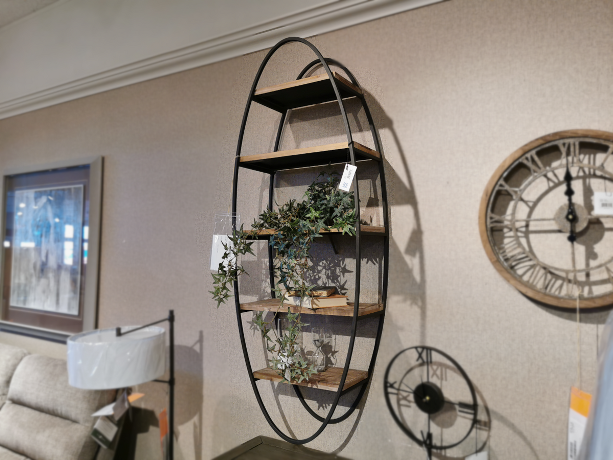 The Mercana 50097 Metort Wall Shelf is an oval wall unit with four wooden shelves, elegantly decorated with plants and a framed picture on the left. It is complemented by two nearby wall clocks for a complete display. The unit securely hangs with keyhole wall mounts, adding a stylish touch to your decor.