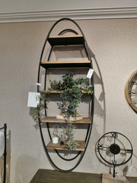 The 50097 Metort Wall Shelf by Mercana is an oval wall unit with keyhole wall mounts and wooden shelves, ideal for displaying books, plants, and a candle. A clock peeks in from the right.