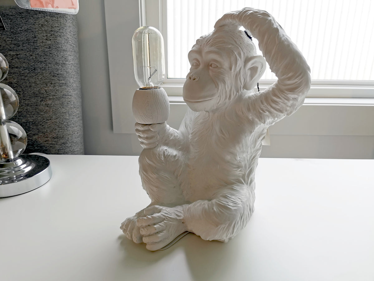 The 65319 Simia Table Lamp by Mercana, a white lamp shaped like a thoughtful monkey holding a light bulb, sits elegantly on a table next to the window.