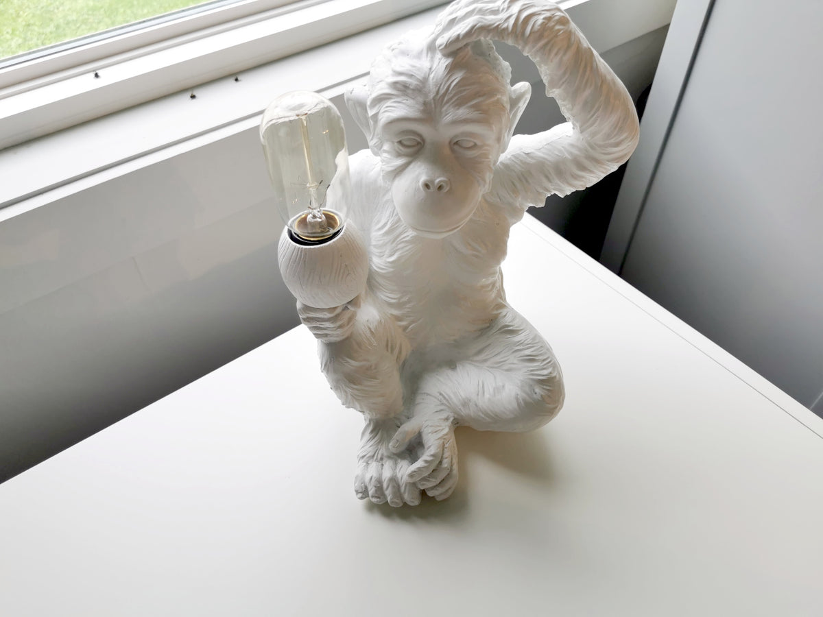 The 65319 Simia Table Lamp by Mercana, featuring a thoughtful monkey figure holding a bulb, rests elegantly on a white surface near the window.