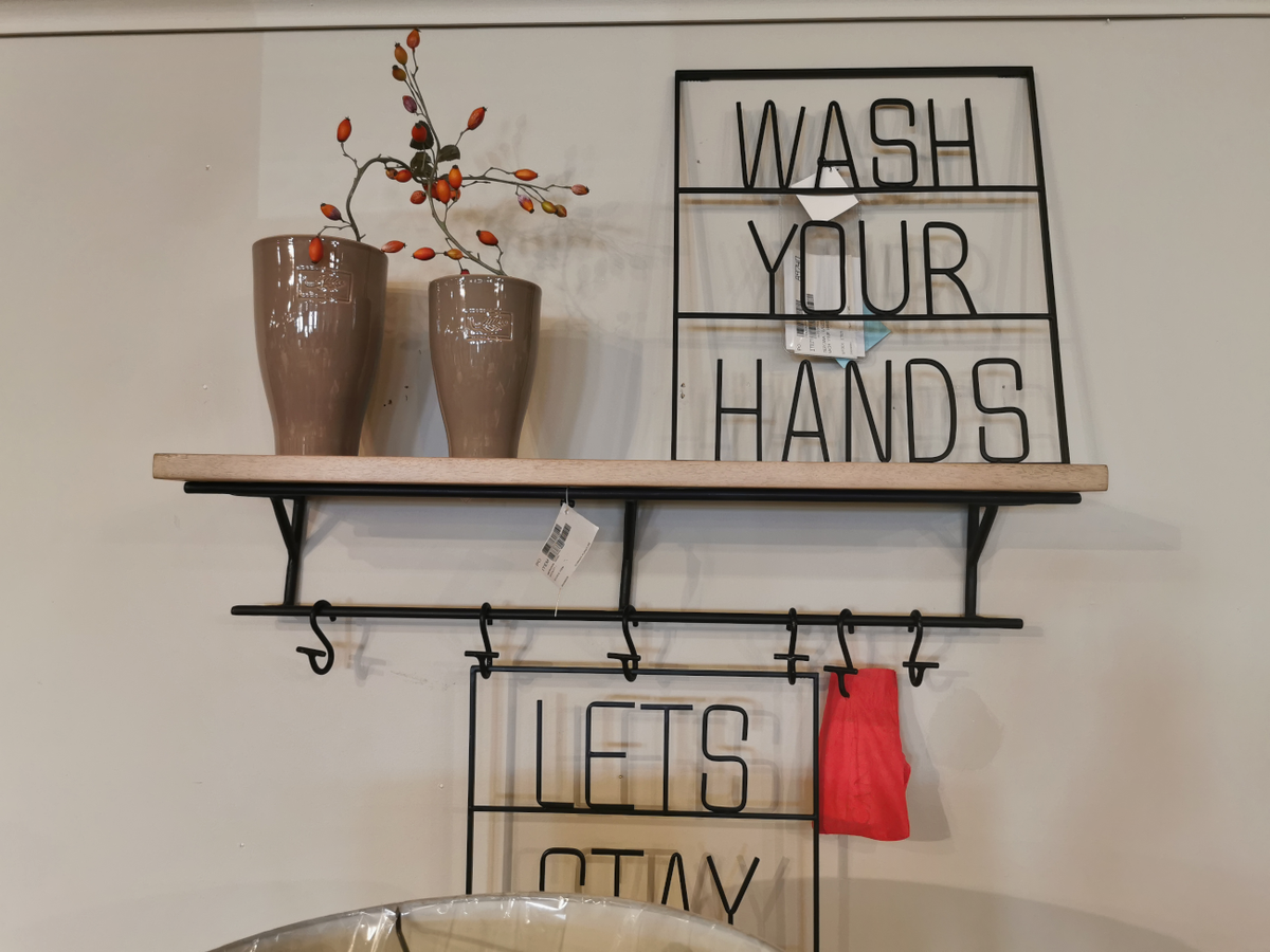 The Mercana 68493 Schutt Shelf showcases a modern design with its wall-mounted Indian mango wood construction, adorned with a "Wash Your Hands" sign, two vases with branches, and hooks holding a vibrant red cloth.