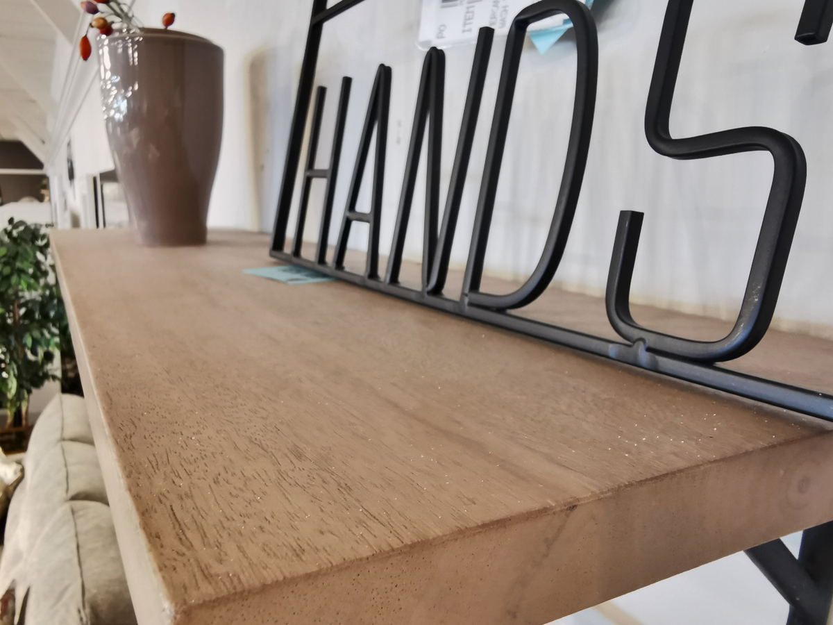 A close-up of the Mercana 68493 Schutt Shelf, crafted from Indian mango wood, showcases a brown vase and black metal letters spelling "HANDS," highlighting its modern design. In the background, a blurred sofa and plant enhance the depth of the scene.