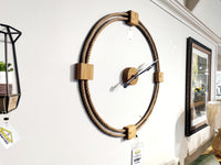 The 68527 Worthing Wall Clock by Mercana showcases an open-face design, highlighted by a rope frame and stylish wooden accents. Its hands, artfully set at 10:10, enhance its visual charm. Price tags dangle from it as it graces a spotless white wall with elegance.