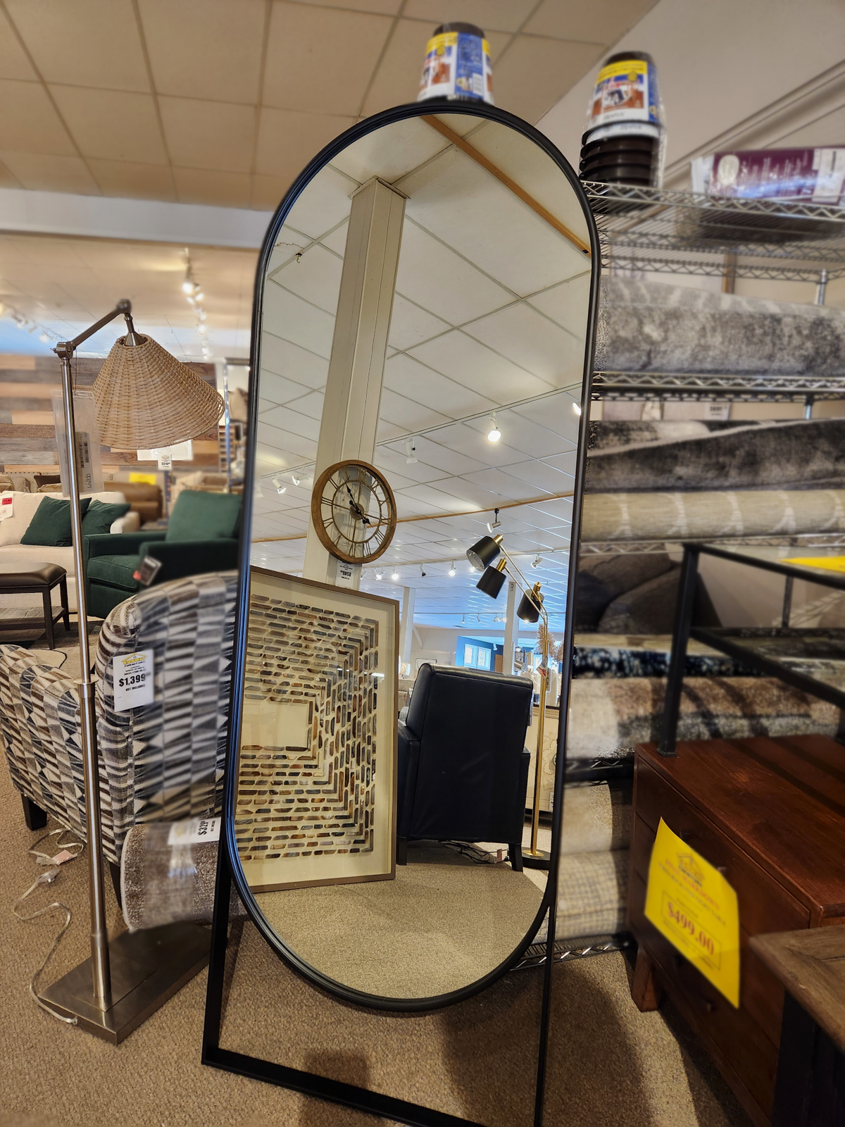 The 69538 Sadie Rounded Floor Mirror by Mercana, featuring a sleek black finish and a sturdy metal frame, is elegantly displayed in a furniture store. It is surrounded by stylish rugs, an eye-catching clock, and a chic lamp—all on sale.