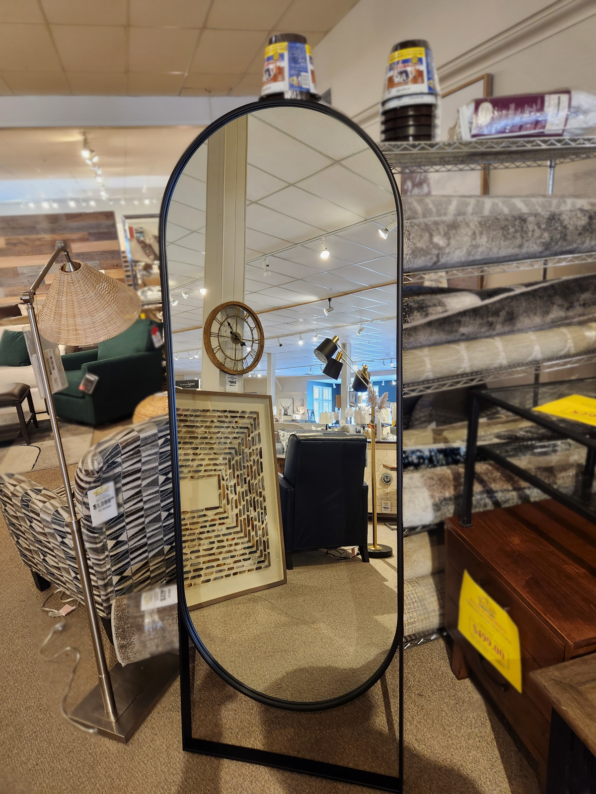 The 69538 Sadie Rounded Floor Mirror by Mercana, featuring a sleek black finish and metal frame, is displayed in a store surrounded by various home decor items and furnishings.