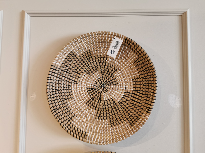 The 69601 Luna by Mercana is a beautifully crafted round basket that serves as an exquisite wall decor piece. It showcases a captivating spiral pattern in black and natural tones, hanging elegantly on the wall as a striking seagrass accent, complete with its price tag.