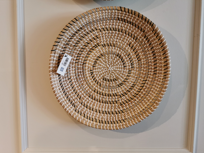 Introducing the 69602 Avel by Mercana, a round woven basket featuring an intricate circular pattern that serves as a chic wall hanging plate against a white wall. This piece is crafted from light brown seagrass and secured with a dark brown string, offering natural hues that bring rustic elegance to any space.