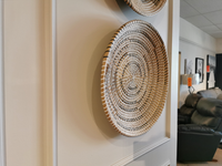 The 69602 Avel wall hanging plate by Mercana, made from light brown seagrass and dark brown string, introduces an earthy touch to your decor. It elegantly complements leather sofas and framed pictures, enhancing the harmonious blend of textures on the white wall.