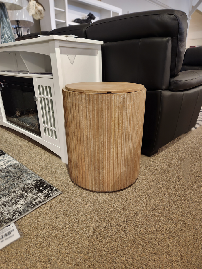 The 69882 Terra Round Side Table by Mercana, crafted from solid mango wood with vertical grooves, rests elegantly on a beige carpet between a black couch and a white cabinet.