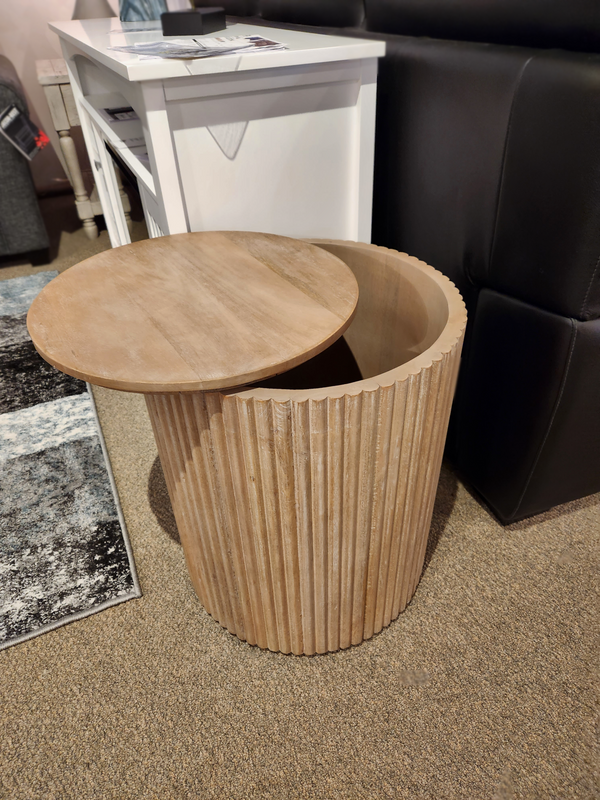 The 69882 Terra Round Side Table by Mercana is crafted from solid mango wood and includes a removable lid that reveals hidden storage. This elegantly round wooden piece complements a chair on a carpeted floor perfectly.