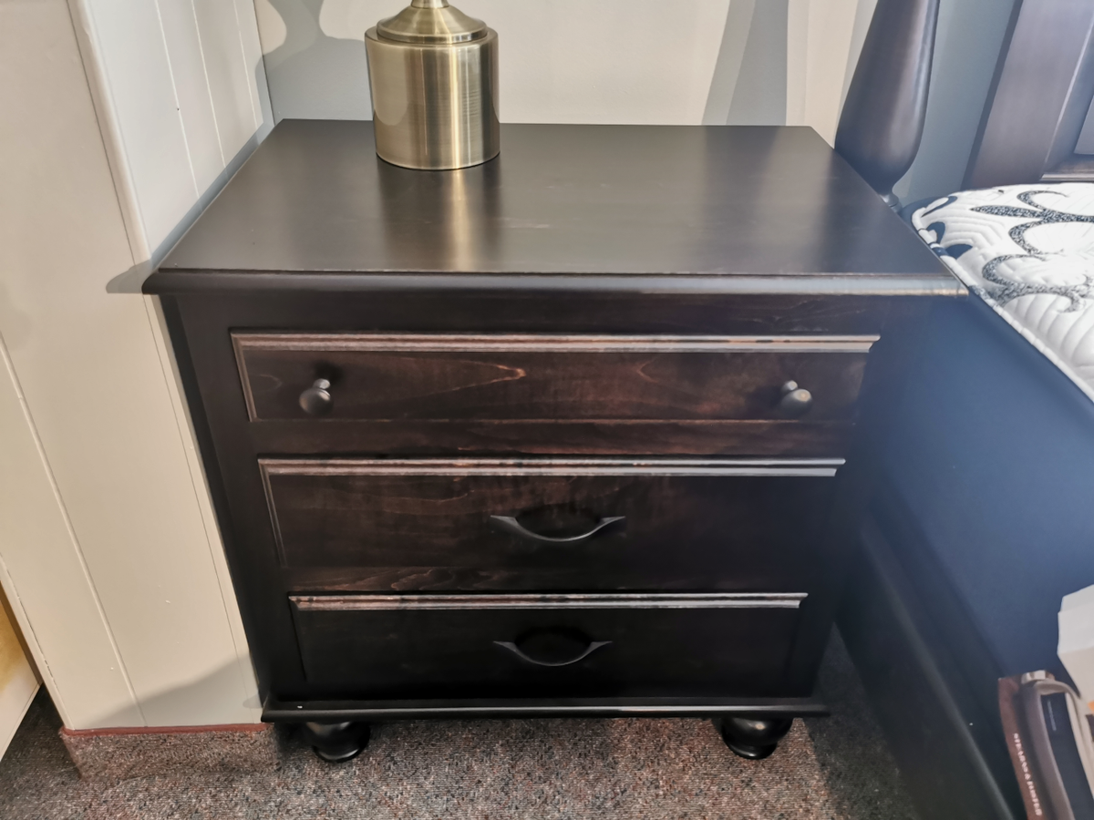 The Muskoka Maple 3 Drawer Nightstand from Munros, featuring a rich dark wood finish, stands elegantly beside the bed. Its three drawers complement the bedroom set beautifully, while a lamp softly illuminates the corner.