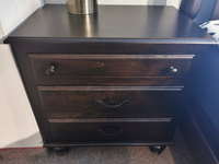 The Muskoka Maple 3 Drawer Nightstand by Munros, with a lamp on top and featuring three drawers with black knobs and handles, complements the Munros Home Furnishings bedroom set. Its rich mahogany stain adds a touch of elegance to any room.