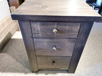 A Munros 3 Drawer Pine Nightstand with round metal knobs stands on a carpeted floor.