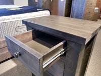 Munros' 3 Drawer Pine Nightstand in a modern showroom, with part of a bed visible in the background.