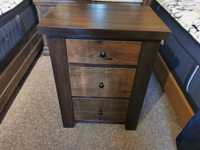 Munros 3 Drawer Pine Nightstand with dark knobs, placed between two beds on a carpeted floor.