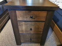 A Munros 3 Drawer Pine Nightstand with black knobs stands on a carpeted floor in a modern bedroom.