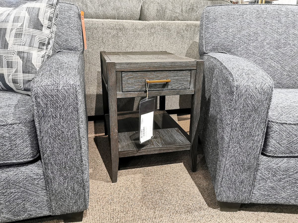 The Null Furniture Manhattan 1022 end table with a drawer and brass hardware sits between two gray upholstered chairs.