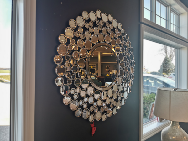 The Mt849 Andromeda by Renwil, featuring a circular mirror adorned with an intricate arrangement of smaller round mirrors, makes for a captivating conversation piece on a dark wall, elegantly reflecting the room and nearby lamp.