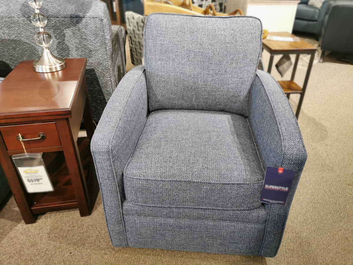 A Superstyle 37-59C Swivel Chair, upholstered in gray for comfortable support, is displayed in a furniture store next to a wooden side table with a lamp.