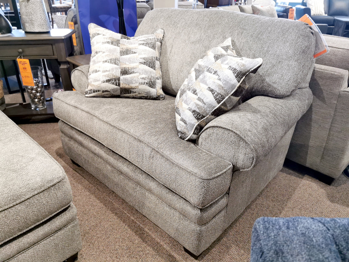 In the furniture showroom, a cozy Trendline 4610 chair and a half sits elegantly on a carpeted floor, accompanied by its matching gray upholstered loveseat adorned with two patterned pillows, showcasing their custom designer fabric.