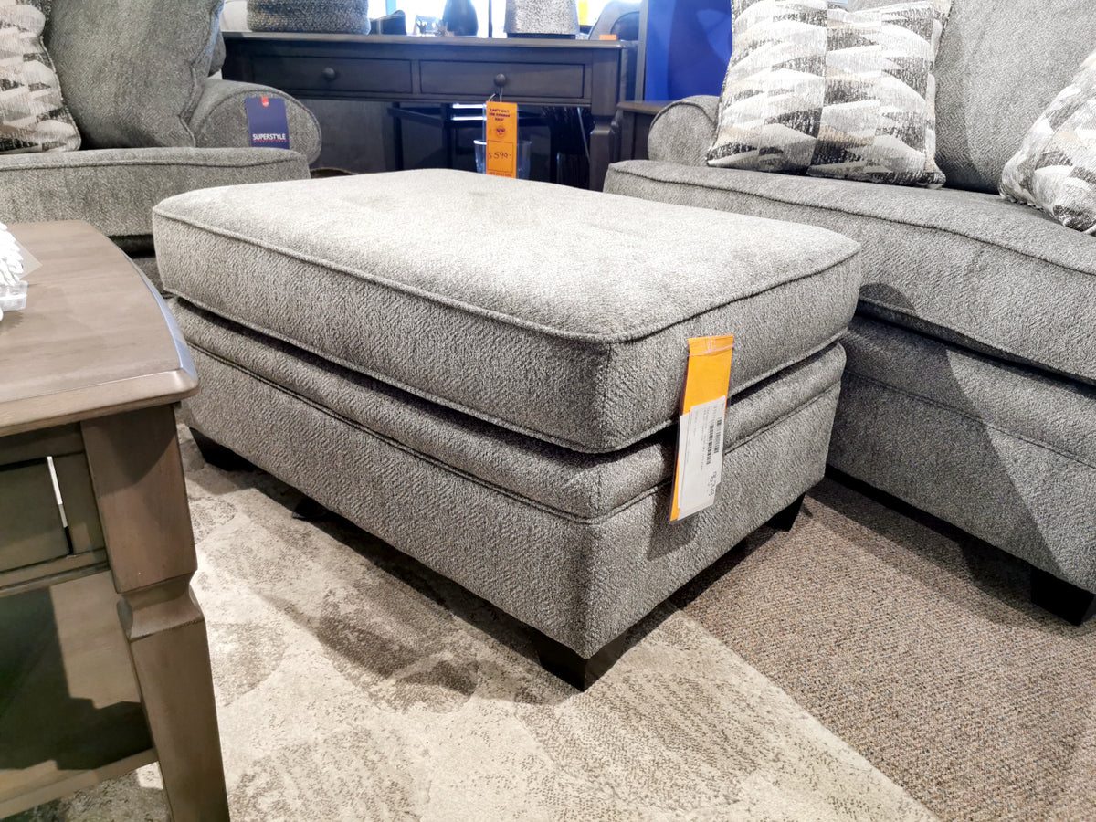 The 4610 Rectangular Ottoman by Superstyle, crafted in Canada and upholstered in gray fabric, is elegantly showcased in a furniture showroom, positioned between a coordinating sofa and a wooden table. With a variety of fabric options at your disposal, this piece offers custom upholstery possibilities to complement any decor.