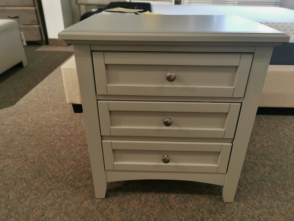 The Vaughan Bassett Bonanza bb26 2-Drawer Nightstand, featuring a gray wooden design with cherry veneers, stands on a carpeted floor in a furniture showroom, making it ideal for adding charm to any transitional space.