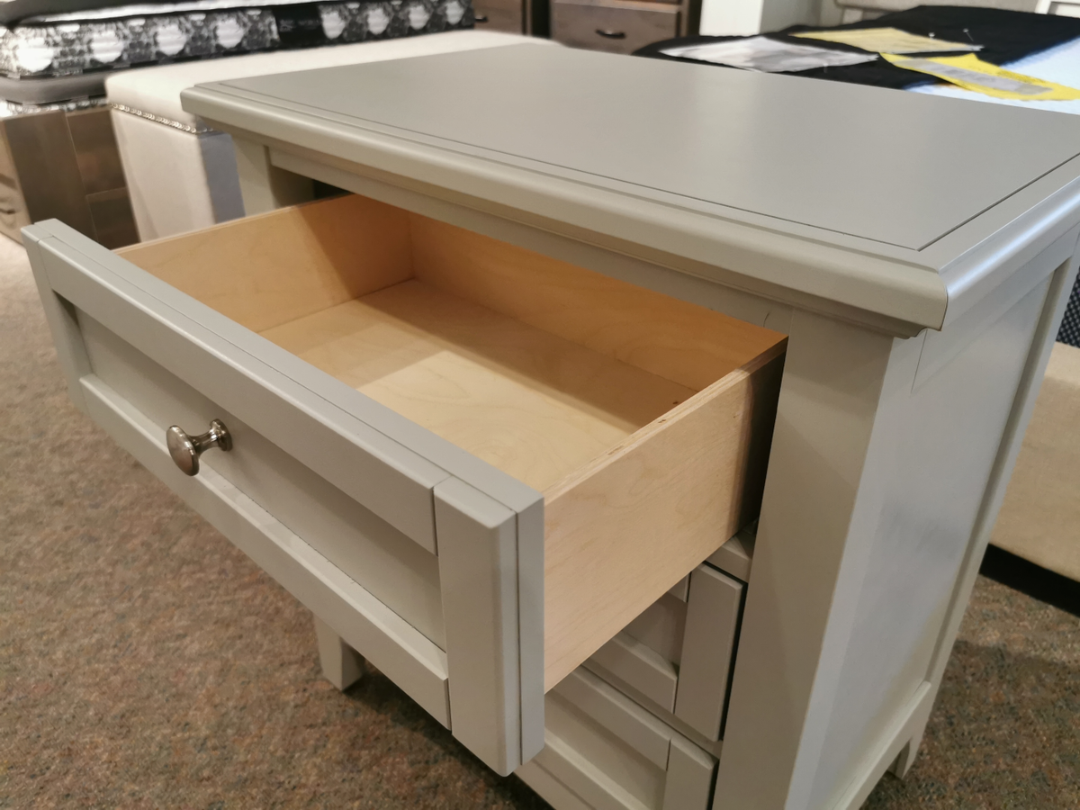 The Bonanza BB26 2-Drawer Nightstand by Vaughan Bassett, featuring a light gray finish with satin nickel hardware, includes an open top drawer that showcases its wooden interior. It is gracefully displayed on a carpeted floor in a furniture showroom.